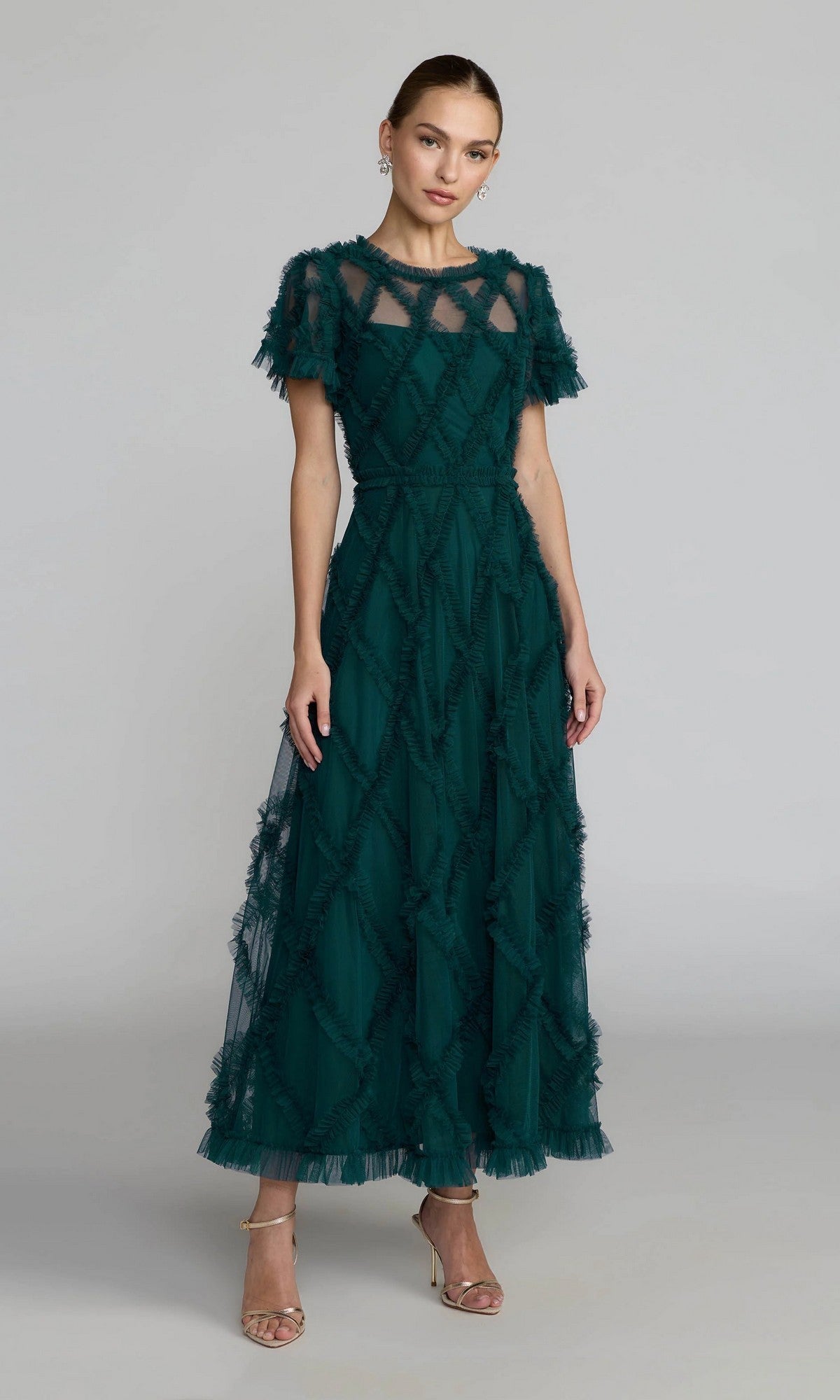A woman poses in a studio wearing the Mac Duggal 8278 Short Sleeve Midi Formal Dress in dark green, featuring a sheer upper bodice and textured pattern, elegantly paired with gold high-heeled sandals.